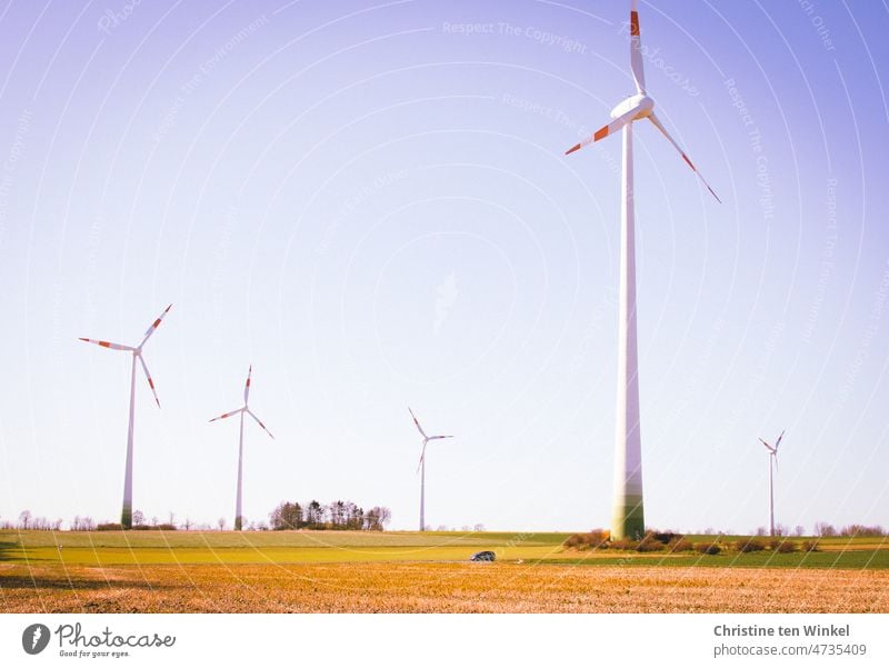 Windkraftanlagen im Sonnenschein, noch unbestellte Felder, Bäume im Hintergrund.  Ein Auto fährt quer durch das Bild Energiekrise Windenergie regenerativ