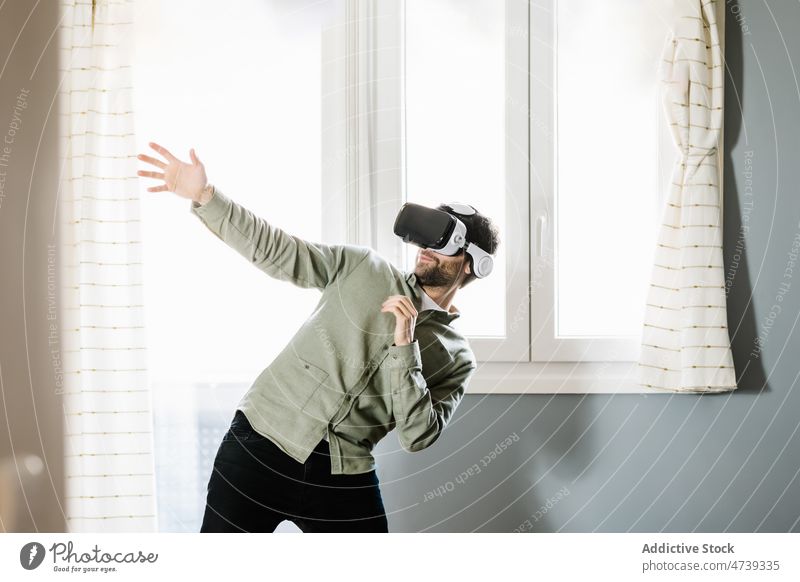 Junger Mann erkundet den Cyberspace mit einer VR-Brille zu Hause Virtuelle Realität Lächeln Erfahrung Innovation gestikulieren simulieren unterhalten digital