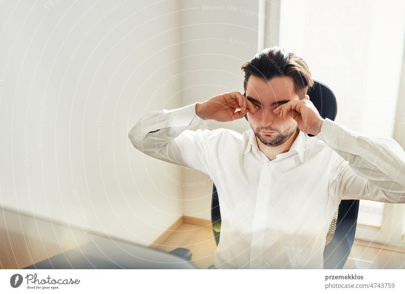 Müder, erschöpfter junger Mann, der sich die Augen reibt, weil er von langer Computerarbeit überanstrengt ist. Mann sitzt am Computerbildschirm Büro arbeiten