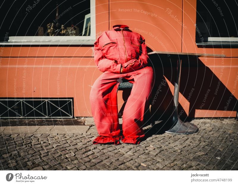 Kopflos im roten Overall sitzt er am schiefen Bistrotisch Latzhose Sitz Prenzlauer Berg Berlin Torso Hausmauer Puppe Kopfsteinpflaster Bekleidung ohne Kopf