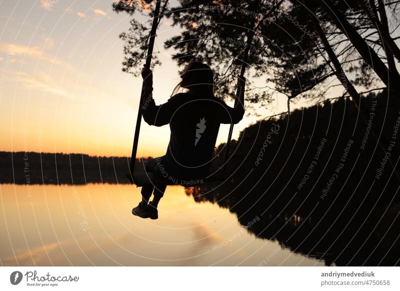 Silhouette einer romantischen jungen Frau auf einer Schaukel über einem See bei Sonnenuntergang. Junges Mädchen Reisende sitzen auf der Schaukel in der schönen Natur, Blick auf den See