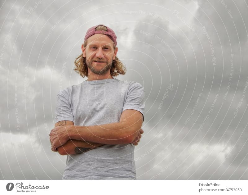 Mann vor Wolkenhimmel Porträt Bart rot grau cappy Wolkendecke verschränkte Arme Locken Lächeln Tattoo blond blaue augen skater