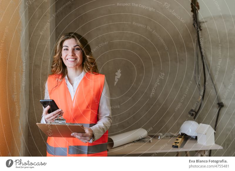 professioneller Architekt Frau auf der Baustelle mit Handy und Blaupausen. Hausrenovierung Arbeitsbereich Schutzhelm Schutzmantel Grundbesitz Kaukasier Glück