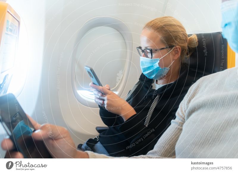 Eine junge Frau trägt eine Gesichtsmaske und benutzt ein Smartphone während einer Reise im Flugzeug. Neue normale Reise nach Covid-19 Pandemie Konzept. reisen