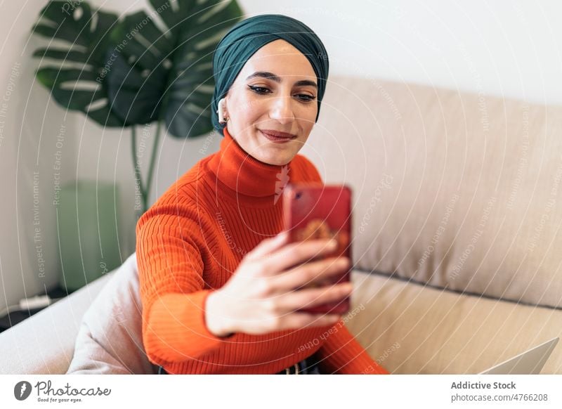 Muslimische Frau macht Selfie zu Hause Porträt heimwärts Wohnzimmer Smartphone Lächeln Sofa Musik zuhören Wochenende muslimisch ethnisch Islam Kopftuch