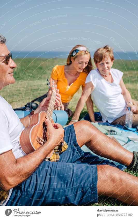 Älterer Mann spielt Ukulele für seine Familie auf einer Decke sitzend während eines Ausflugs Senior Spielen Musical Instrument im Freien Erwachsener