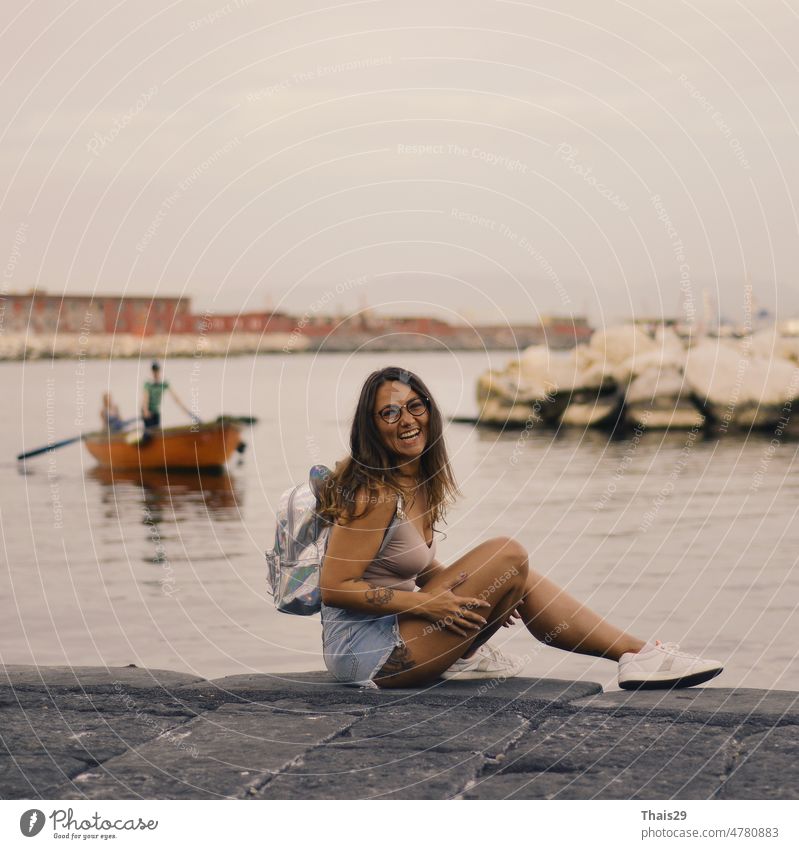 Junge hübsche Frau sitzt allein auf dem Pier in der Nähe des Meeres, lächelnd und lachend Rücken Hintergrund Strand schön Windstille Großstadt Abend Freiheit