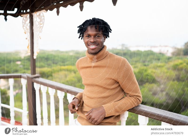 Glücklicher schwarzer Mann mit Dreadlocks auf Terrasse Rastalocken heiter Stil Vorschein Porträt Outfit Balkon Rollkragenpulli Landschaft Zaun ländlich hölzern