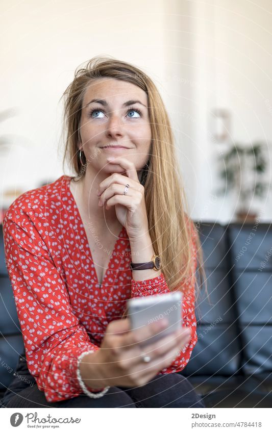 Porträt einer einzelnen schönen kaukasischen Frau, die ein Handy in der Hand hält und auf dem Sofa sitzt, während sie nachdenklich wegschaut Denken Smartphone