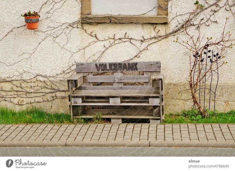 eine aus Europaletten gebaute Holzbank mit der Aufschrift  VOLKSBANK  steht vor einem Haus auf der Wiese neben dem gepflasterten Fußweg Bank Volksbank Humor