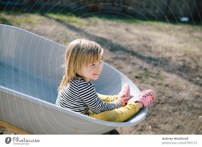 Kleines Mädchen, das Spaß daran hat, in einer Schubkarre durch den Hinterhof zu fahren Ackerbau blond Karre Kaukasier Kind Kindheit niedlich Gerät Gesicht