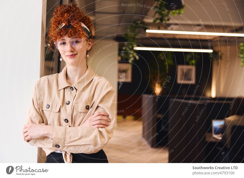 Porträt einer selbstbewussten jungen weißen Frau mit roten lockigen Haaren, die eine Brille trägt, lächelt und in die Kamera schaut, in einem Coworking Space