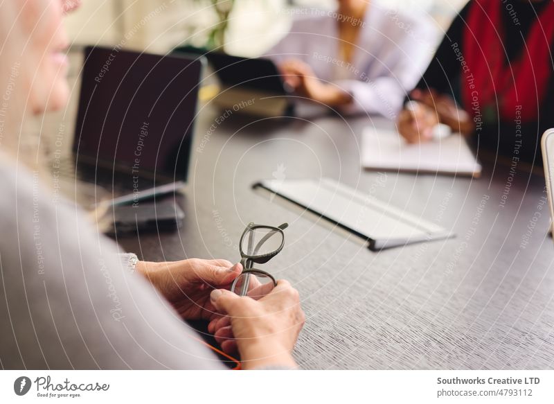 Nahaufnahme einer Frau, die eine Brille hält und sich mit den Händen auf einen Besprechungstisch stützt, mit weiblichen Kollegen im Hintergrund Geschäftsfrau