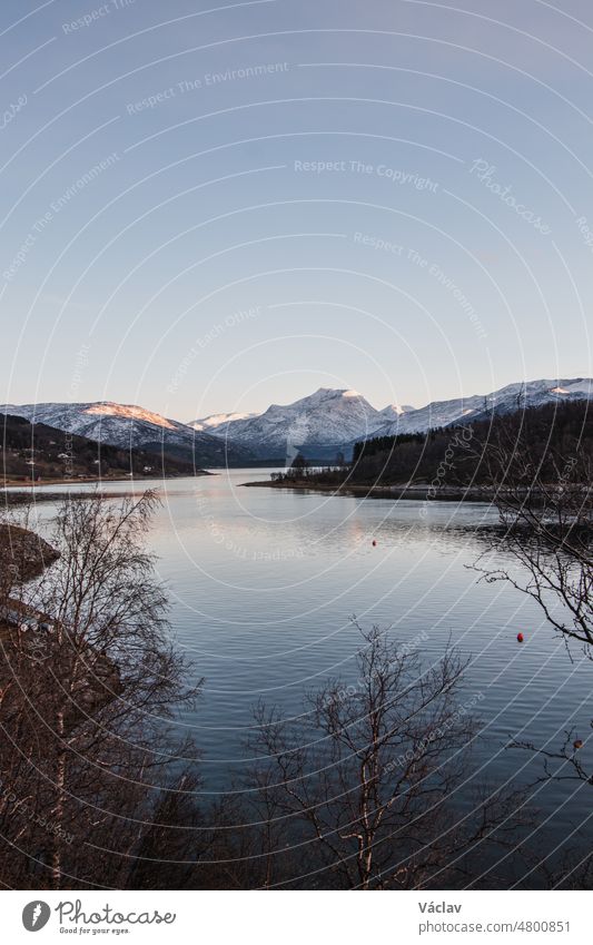 Verschneite Berghügel mit Blick auf den Rombakensee in der Nähe der Stadt Narvik in Nordnorwegen, Skandinavien. Die letzten Splitter des Sonnenuntergangs fallen auf die weißen Berggipfel.