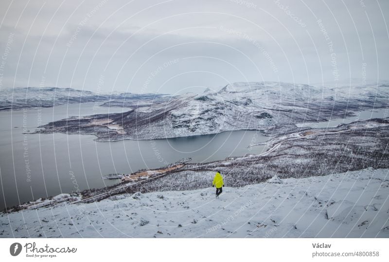 Frostiger Sonnenaufgang auf dem Berg Saana in Lappland, Nordwestfinnland. Mann in grüner Jacke geht mit Blick auf den Ylinen Kilpisjarvi und den Rest der kargen Landschaft. Dorf Kilpisjarvi. Grenze zu Norwegen