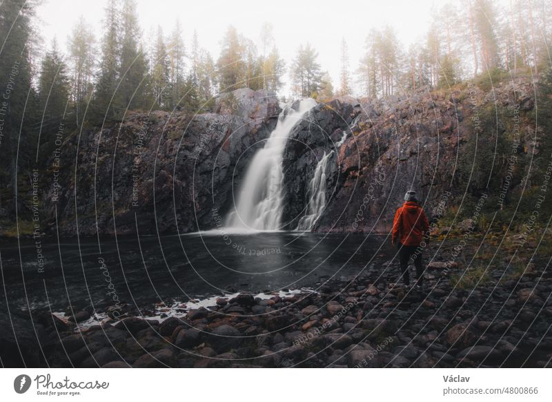 Finnlands berühmter Hepokongas-Wasserfall bei Sonnenaufgang und Nebel, der ins Tal fällt. Touristischer Ort in der Region Kainuu, Finnland. Ein Forscher in einer roten Jacke genießt den Blick auf das Wasser, das vom Felsen fällt