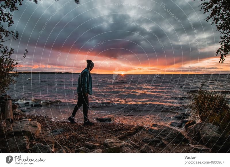 Dramatischer Himmel mit einer Mischung aus Rot, Orange und Violett über den Wellen des Oulujarvi-Sees mit Blick auf einen Mann in blauem Hemd und Mütze, der dieses Juwel am Strand von Kajaani, Region Kainuu, Finnland, beobachtet