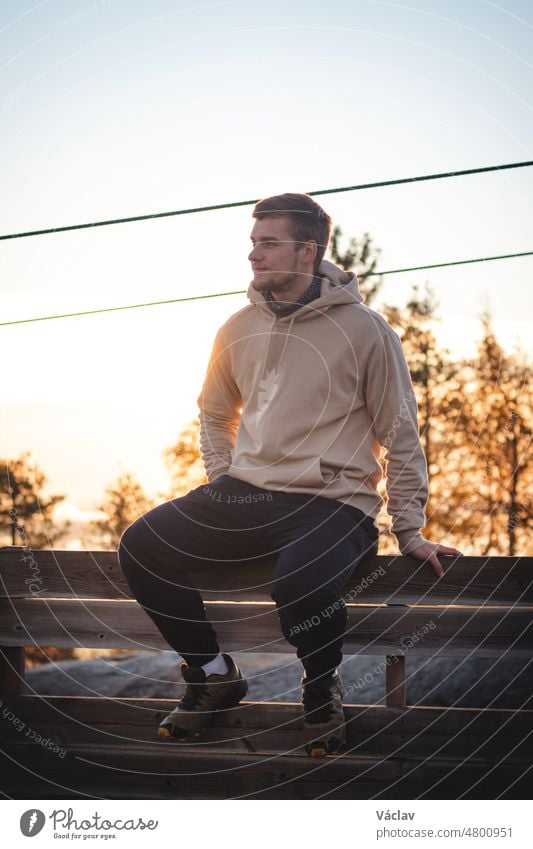 Männliches Modell in einem beigen Sweatshirt sitzt auf dem Gipfel des Berges Vuokatinvaara in der Region Kainuu, Finnland. Ein Mann mit athletischer Figur und einem realistischen Lächeln genießt den Sonnenaufgang und den Herbst