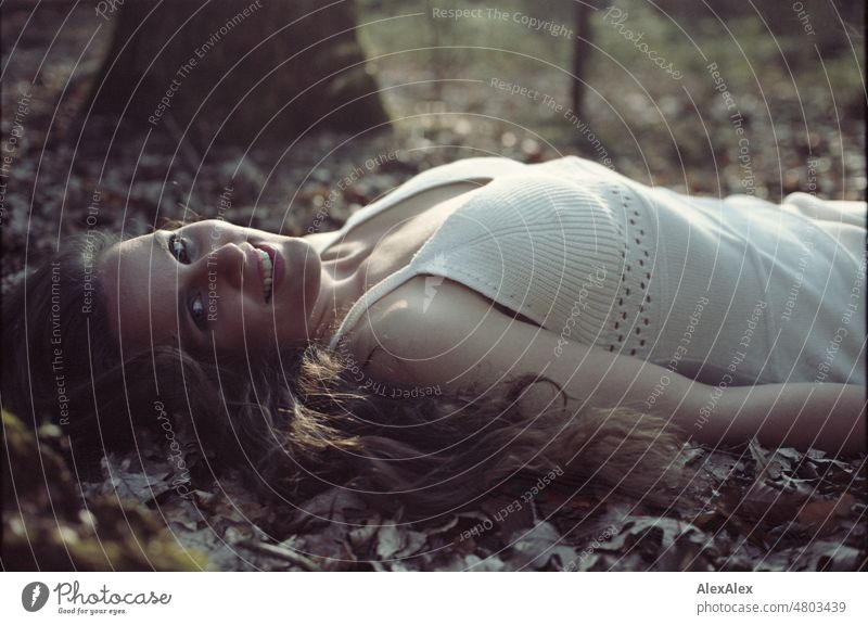 analoges Kleinbild-Portrait einer jungen Frau in weißem Kleid, die in einem Wald im Laub liegt und in die Kamera lächelt junge Frau schön feminin weiblich