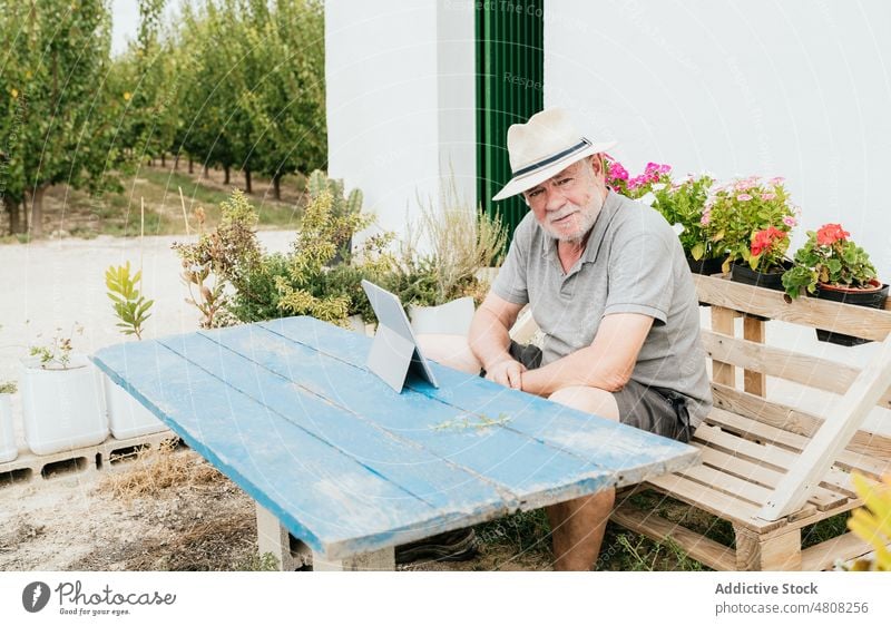 Älterer Bauer benutzt Tablette im Garten Mann Landwirt benutzend Pause Haus Daten Landschaft Sommer männlich Bauernhof Tisch Bank schäbig ruhen Gerät Apparatur