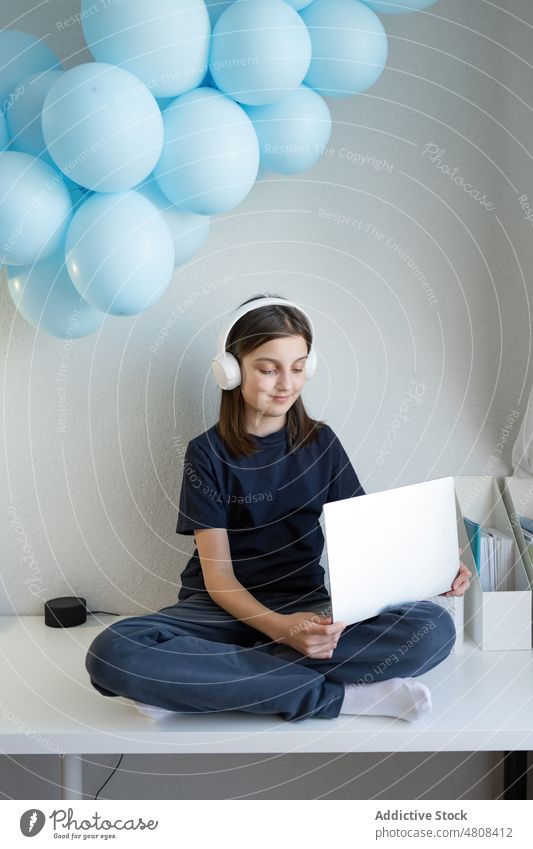 Lächelndes Teenager-Mädchen mit Kopfhörern, das auf einem Tisch sitzt und einen Laptop benutzt benutzend Internet zuschauen Video positiv Beine gekreuzt