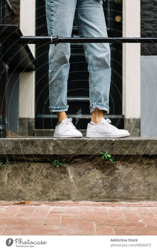 Crop-Dame in modischen Jeans steht vor der Haustür auf der Straße Frau Jeanshose trendy Turnschuh Stil Türschwelle Großstadt Jeansstoff Bein Gebäude Eingang