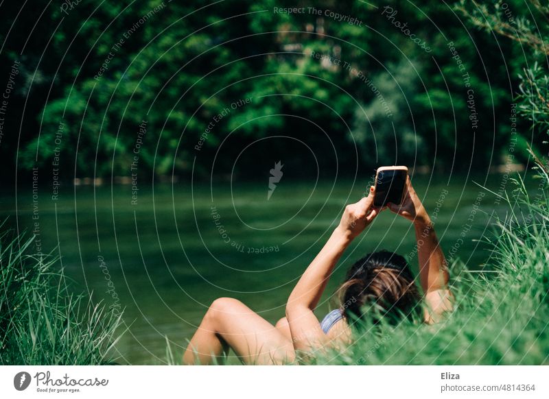 Frau liegt im Grünen am See/Fluss in der Sonne und ist mit ihrem Handy beschäftigt Natur Bikini sonnen Baden Wasser Sommer liegen Erholung Mensch Mädchen lesen