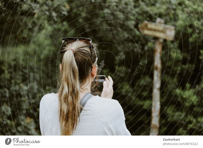 Rückenansicht einer Frau im weißen Oberteil mit Pferdeschwanz, die auf ihr Handy schaut, vor einem Wegweiser Kopf Orientierung suchen Richtung Pfeil Navigation