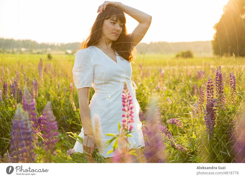 Schöne Frau im weißen Sommerkleid genießt die sommerliche Natur. Pflücken bunte Blumen, atmen frische Luft und Blumenduft, zu Fuß in der sonnigen Feld von Lupinen. Glück Konzept