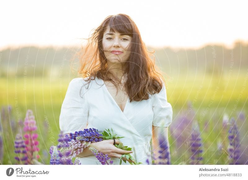 Schöne Frau im weißen Sommerkleid genießt die sommerliche Natur. Pflücken bunte Blumen, atmen frische Luft und Blumenduft, zu Fuß in sonnigen Feld von Lupinen, Wind in den Haaren. Glück Konzept