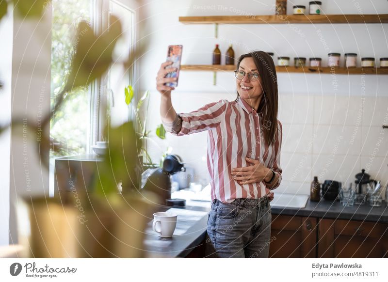 Junge Frau benutzt ihr Smartphone, während sie in ihrer Küche zu Hause steht Handy Technik & Technologie online Internet per Telefon Mitteilung häusliches Leben