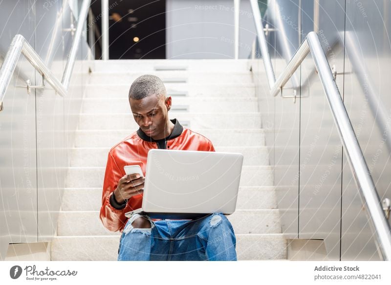 Selbstständiger schwarzer Mann mit Smartphone und Laptop auf der Treppe freiberuflich abgelegen Straße Großstadt multitask männlich jung Afroamerikaner ethnisch