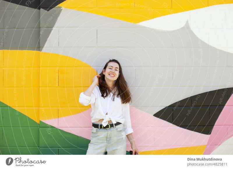 Junge Frau in weißem Hemd und hellblauen Jeans stehen in der Nähe von bunten lebendigen Wand. Candid Lifestyle Sommer Stil Porträt. schön Großstadt Glück