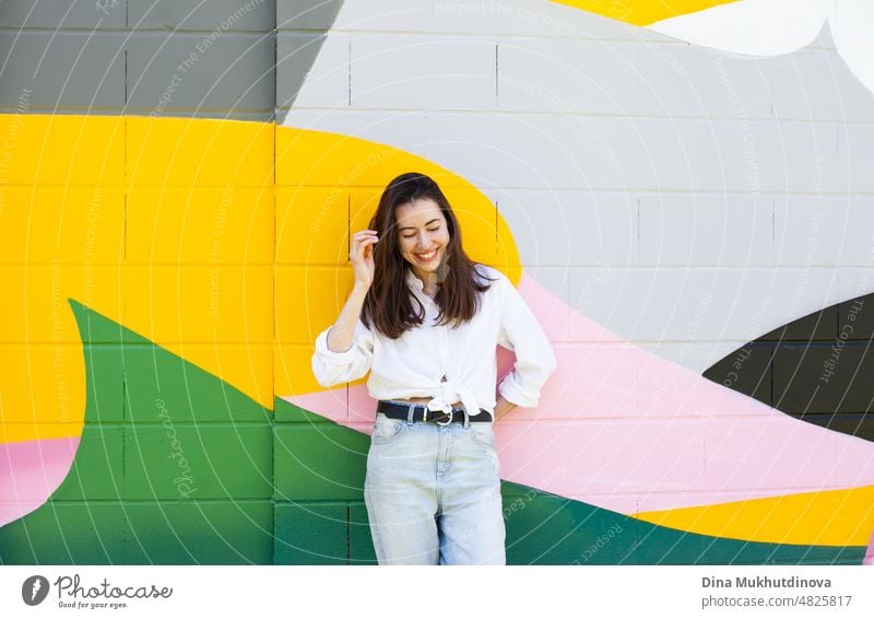 Junge Frau in weißem Hemd und hellblauen Jeans stehen in der Nähe von bunten lebendigen Wand. Candid Lifestyle Sommer Stil Porträt. schön Großstadt Glück