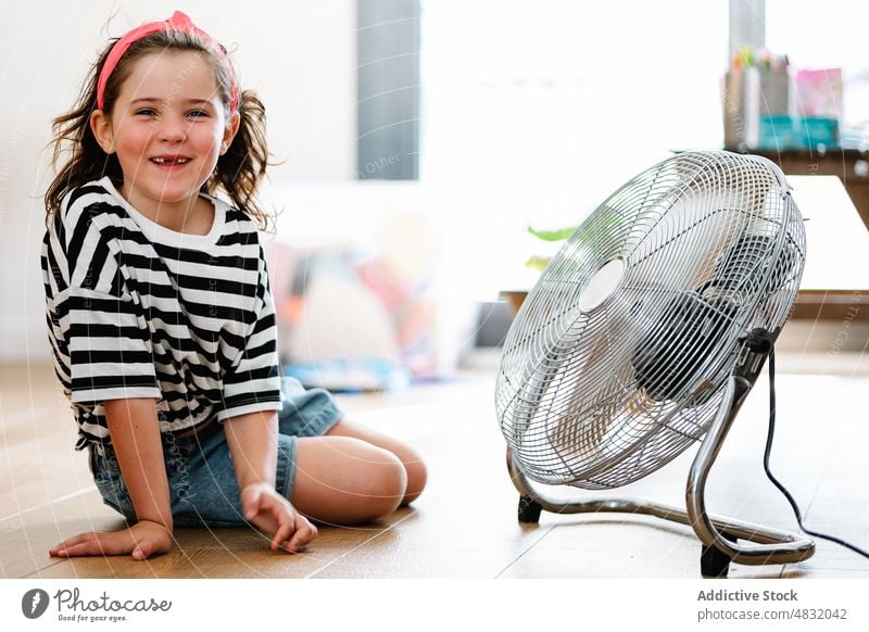 Kleines Mädchen zu Hause auf dem Boden sitzend und mit einem Fächer spielend Ventilator Schlag Spiel jung genießend blasend Spielen Ausdruck Spaß Bewegung Frau