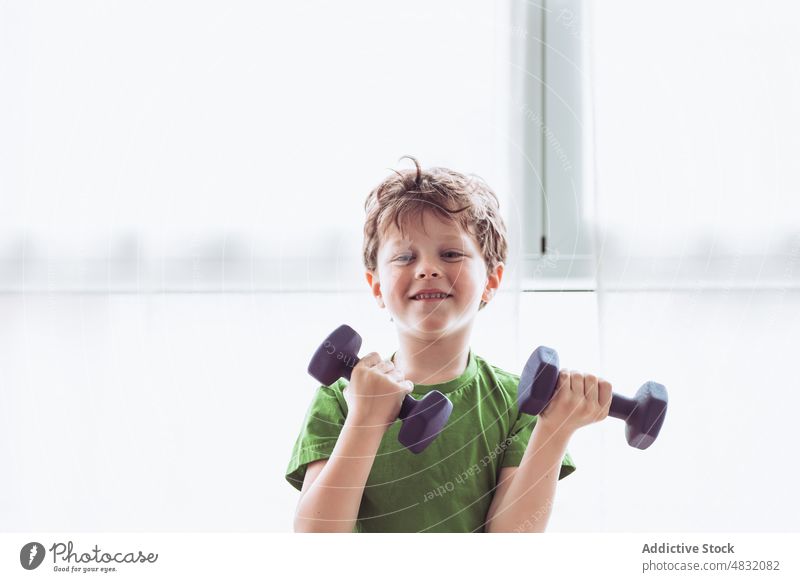 Fröhlicher Junge beim Training mit Hanteln Übung Kurzhantel Fitness Lächeln heimwärts Morgen Glück gymnastisch Kind Fenster Gesunder Lebensstil Sportkleidung