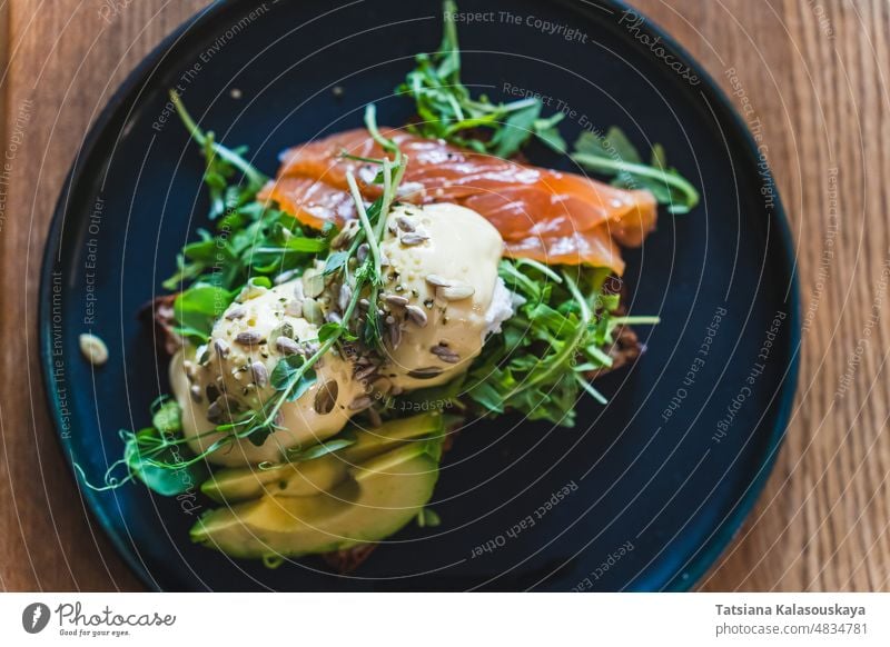 Egg Benedict mit Rucola, Lachs und Avocado auf einem dunklen Teller im Restaurant zum Frühstück Zuprosten Benedikt Brot pochiert Ei Mittagessen grün Gesundheit