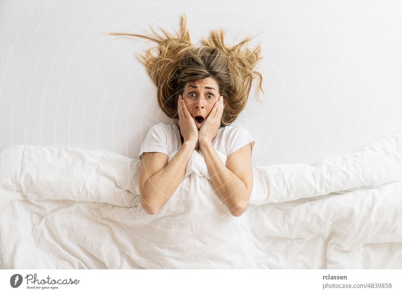 Draufsicht auf eine schockierte Frau, die in die Kamera schaut, während sie im Bett unter einer Decke liegt, mit Platz für Ihren individuellen Text. Top lustig