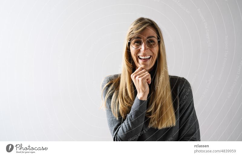 freundliche Frau mit einem schönen Sinn für Humor, die in einem weißen Raum steht, die Hand unter dem Kinn hält, die Wand mit dem Kopierraum zu Hause und in die Kamera schaut