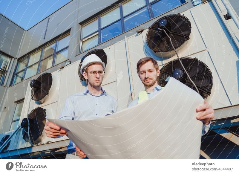 Servicetechniker und Praktikant in Uniform mit Papierzeichnungen an einer industriellen Kühlmaschine Ingenieur Dienst Konstruktion hvac Ventilator Praktikum