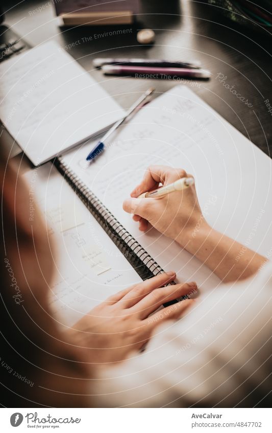 Draufsicht auf den Rücken einer jungen Frau, die zu Hause am Schreibtisch studiert und arbeitet, Hausaufgaben macht, sich mit dem Lehrbuch auf die Prüfung vorbereitet und Notizen macht, selektiver Fokus auf den Bleistift.