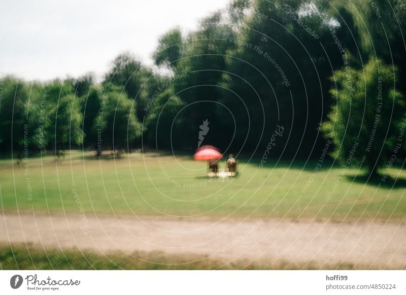 verschwommene Ansicht von zwei Menschen auf Stühlen mit Sonnenschirm im  Park vibrierend ICM-Technik zwei personen pärchen Erholung rosa Schirm