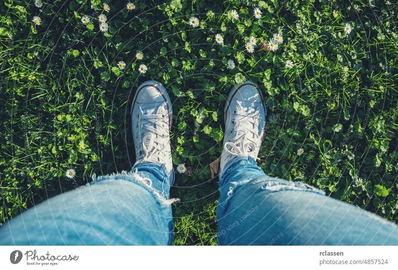 Junge Frau steht in Turnschuhen auf einer Gänseblümchenwiese im Frühling, Hochwinkel-Fußbild oder Flortrait, persönliche Perspektive von oben. pov Menschen