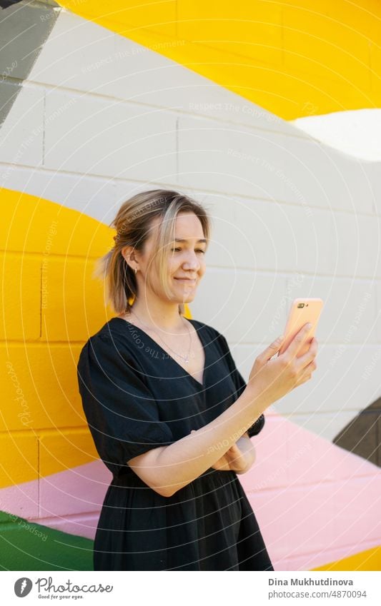 Junge Frau mit Handy steht in der Nähe von bunten Wand nehmen Selfie oder Video-Chat auf dem Smartphone. Bildschirmzeit. Lebensstil mit Technologie. Junge Remote-Arbeiter Mitarbeiter in modernen Büroraum.