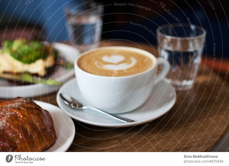 Eine Tasse Kaffee mit Milch auf dem Holztisch mit Gebäck, Glas Wasser und Avocado-Toast. Cappuccino in weißer Tasse Nahaufnahme, Brunch mit Freunden. Candid Lifestyle mit Essen und Trinken. Millennial way of living.