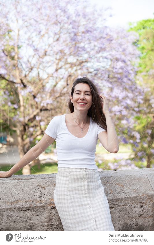 Junge weibliche Brünette Tourist posiert in der Nähe Jacaranda Baum, lächelnd. Neutrale Farben in der Kleidung Stil. Beige Farbe Wände. Frau Tourist trägt weißes T-Shirt.