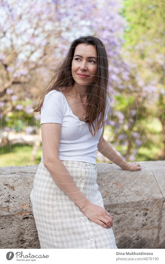 Junge weibliche Brünette Tourist posiert in der Nähe Jacaranda Baum. Neutrale Farben in der Kleidung Stil. Beige Farbe Wände. Frau Tourist trägt weißes T-Shirt.