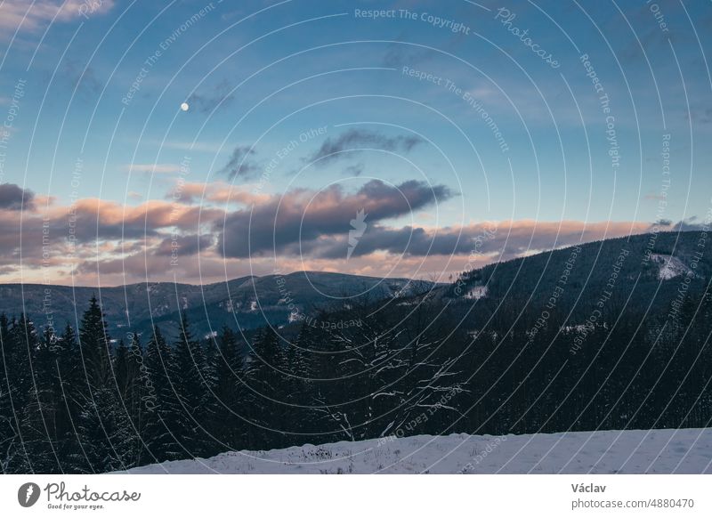 Magischer Abend in den Beskiden im Osten der Tschechischen Republik. Kleine rötliche Wolken strömen auf den Mond zu, der die weiße Landschaft beleuchtet