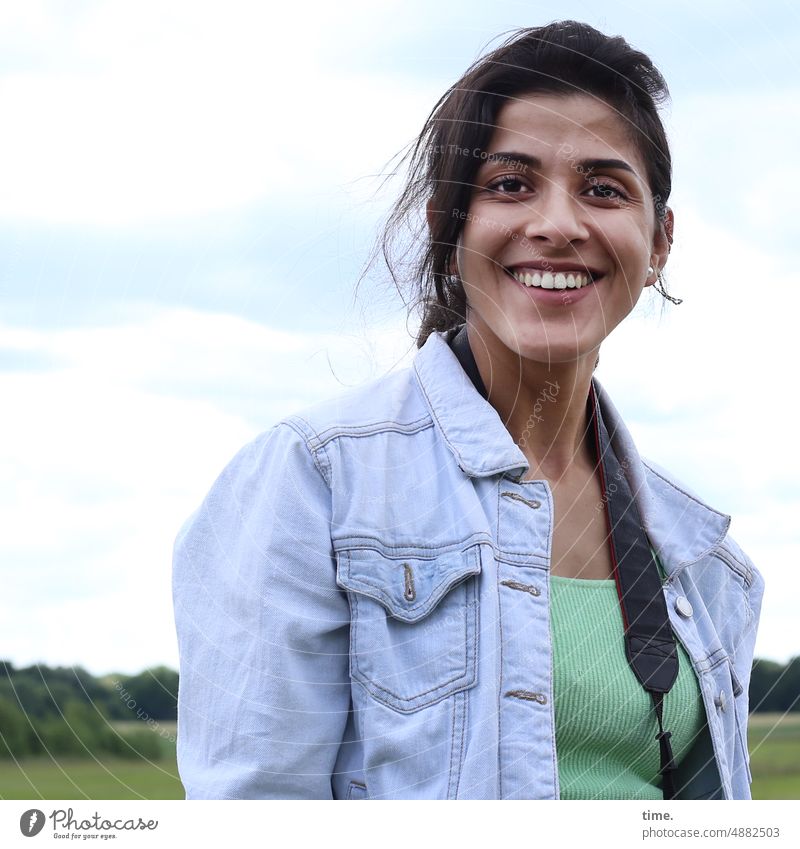 Frau vor Landschaft unter weitem Himmel, gutgelaunt frau weiblich feminin lachen direkt portrait langhaarig dunkelhaarig jacke landschaft kameragurt