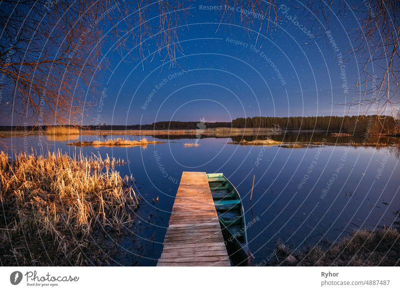 Weißrussland, Osteuropa. Real Night Sky Sterne über alten Pier mit vertäuten hölzernen Fischerboot. Natürliche Sternenhimmel und Landschaft auf dem Land mit See-Fluss im frühen Frühling Nacht. Russische Natur